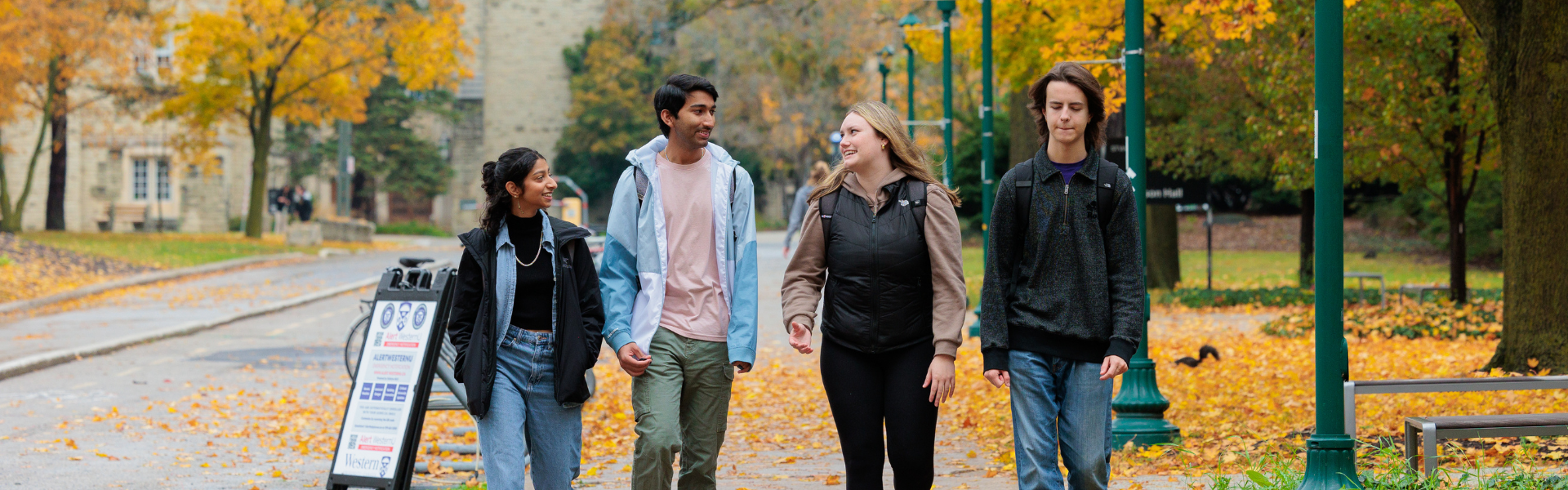 Four people walk along a tree-lined campus path in autumn, surrounded by vibrant yellow and orange foliage and fallen leaves