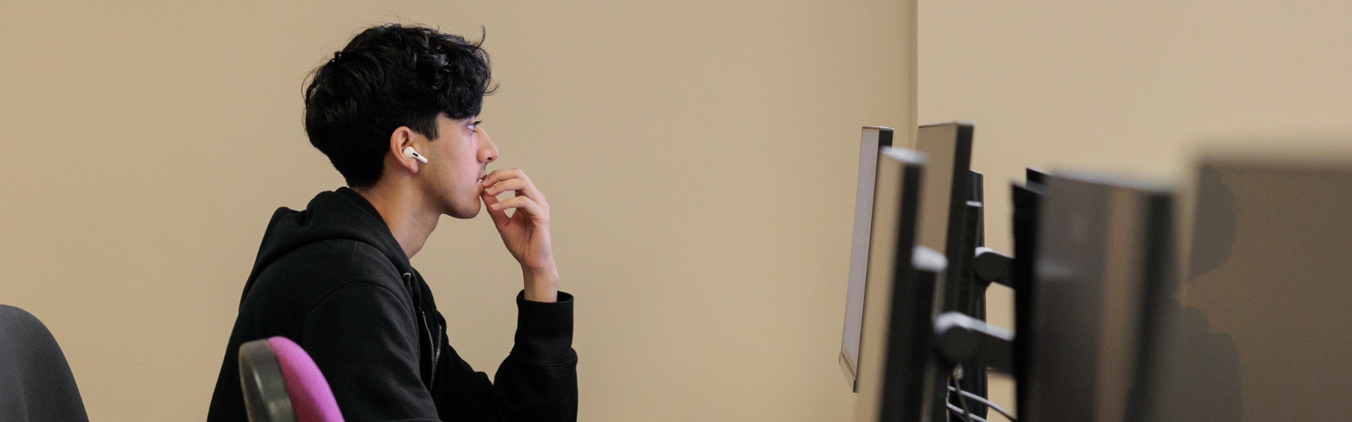 Focused individual in black hoodie and wireless earbuds working at a desk with multiple computer monitors, in a minimalist beige office setting