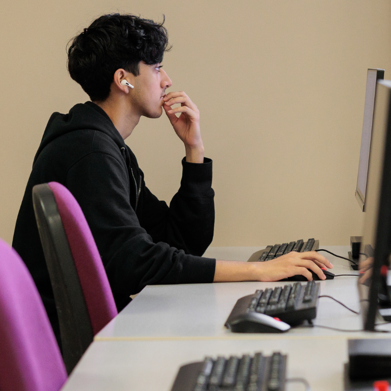 Focused individual in black hoodie and wireless earbuds working at a desk with multiple computer monitors, in a minimalist beige office setting