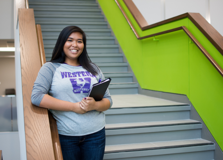 A smiling student in a Western Mustangs shirt holds a notebook while standing on a modern staircase