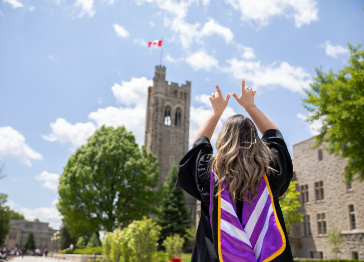 Graduate in gown facing a stone building with a Canadian flag, raising hands in celebration.
