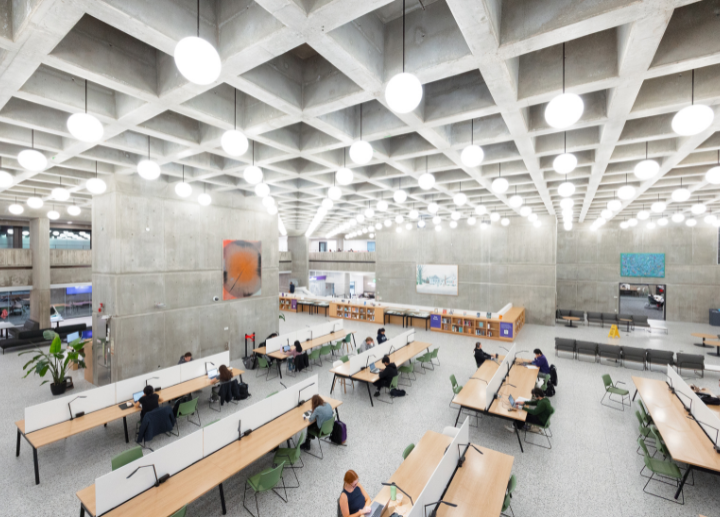 Large modern study hall with concrete walls, grid ceiling lights, long tables, and people working on laptops