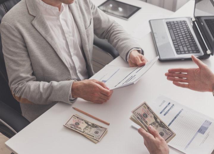 Two people seated at a desk in an office setting, exchanging cash and reviewing documents, with a laptop, papers, and a pencil on the des