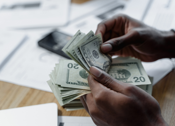 Hands counting a stack of U.S. twenty-dollar bills on a wooden table, with additional cash, papers, and a smartphone in the background