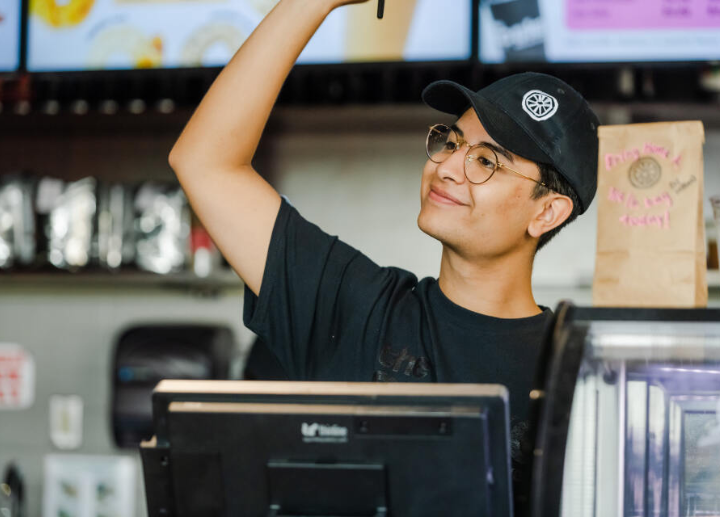 A student working at the Spoke standing at a checkout, waving for the next person in line to come up to the till